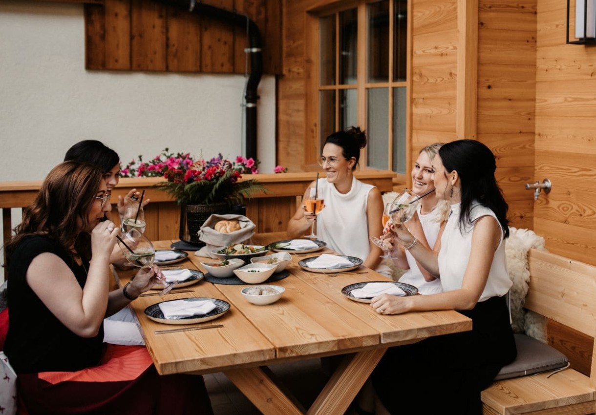 Fünf Frauen sitzen fröhlich um einen Holztisch und genießen Getränke