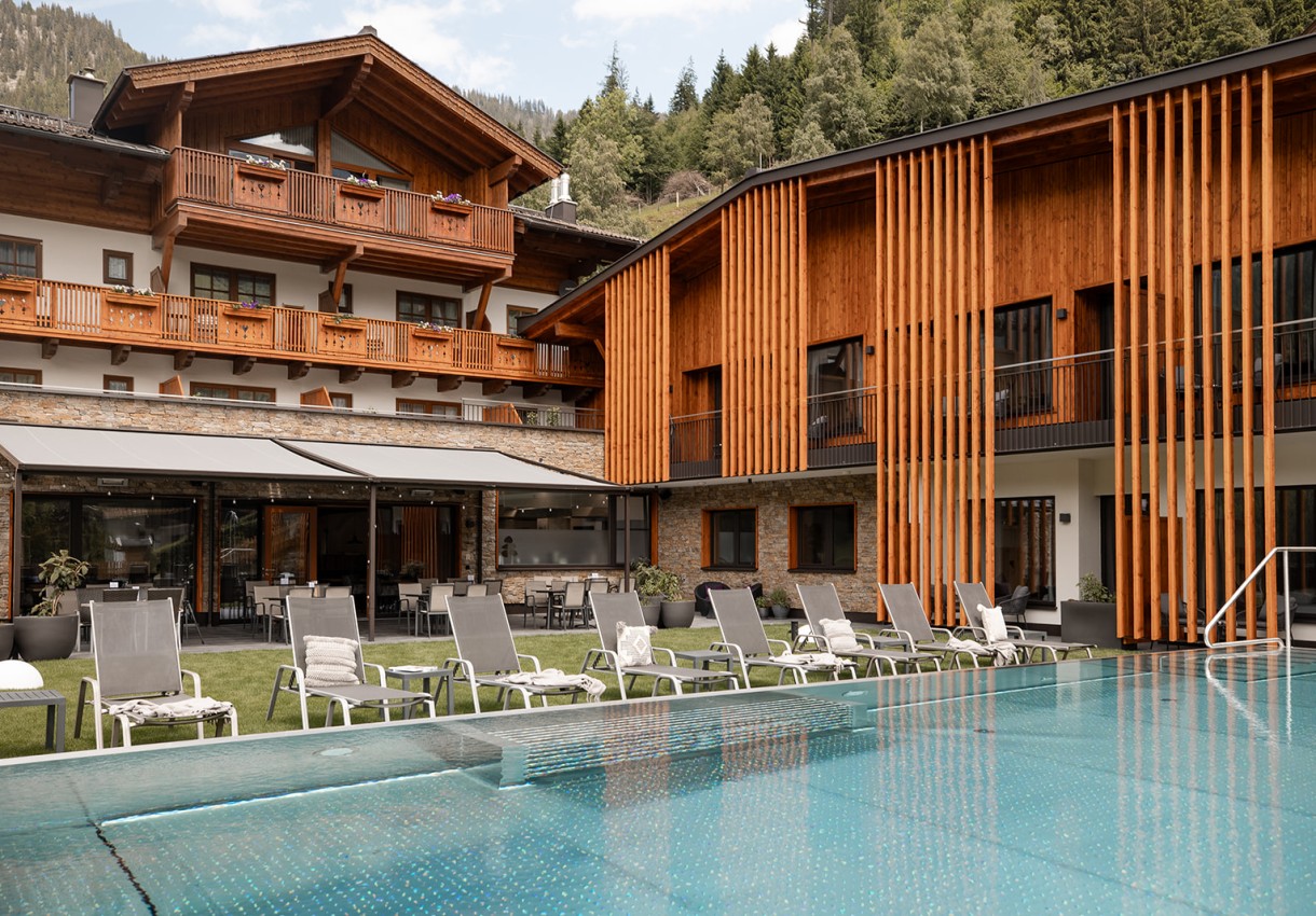Hotel Dorfer mit Holzfassade, Pool und Liegestühlen vor bewaldeten Hügeln
