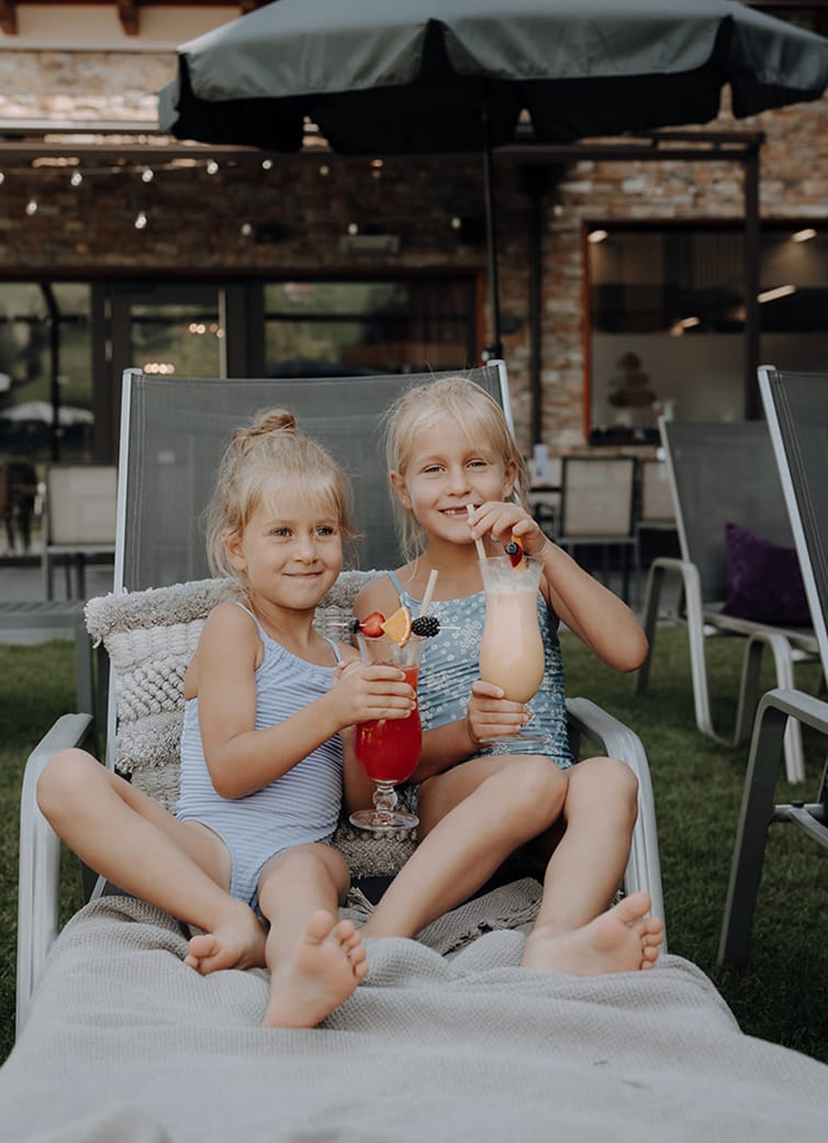 Zwei M&auml;dchen in Badeanz&uuml;gen trinken bunte Cocktails auf einer Liege im Garten