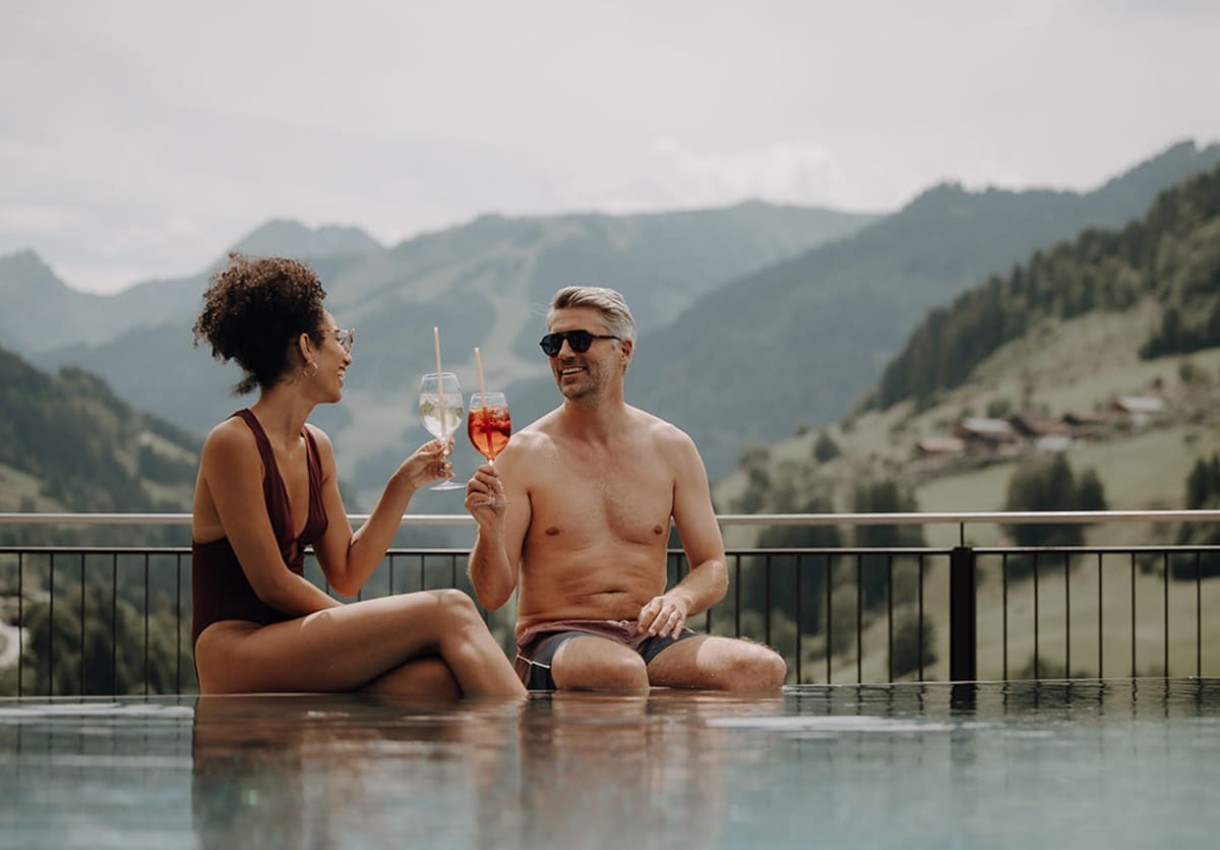 Ein Paar sitzt am Pool und st&ouml;&szlig;t mit Getr&auml;nken an, vor einer Berglandschaft