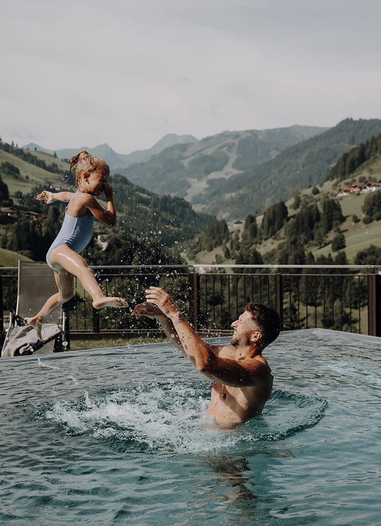 Kind springt lachend in die Arme ihres Papas im Pool mit Bergblick
