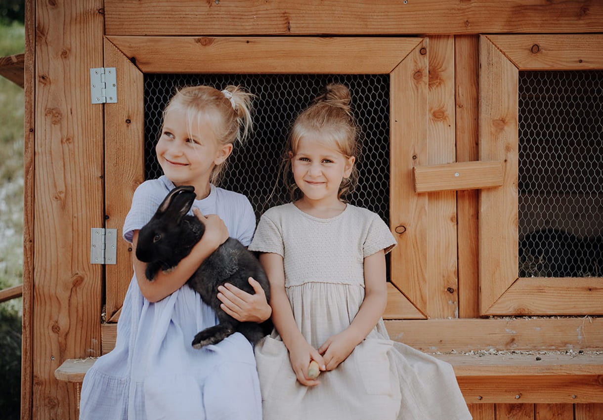 Zwei M&auml;dchen sitzen vor einem Hasenstall mit Kaninchen im Arm