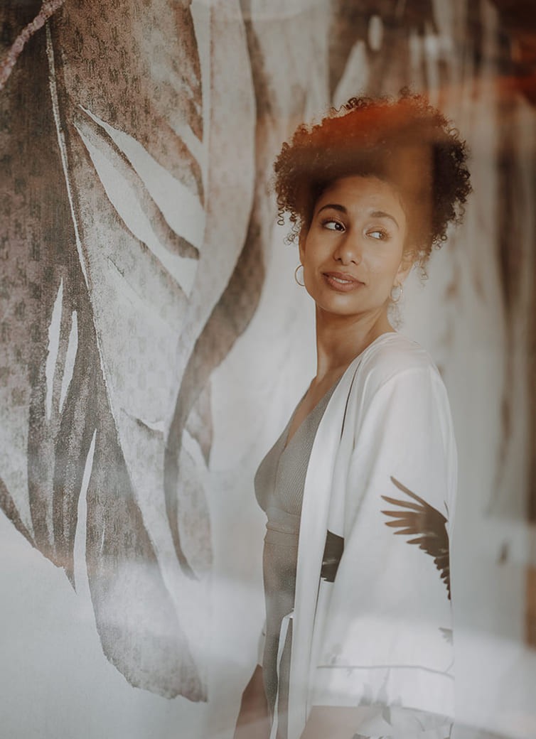 Frau mit lockigem Haar l&auml;chelt vor einer Wand mit Bl&auml;ttermuster