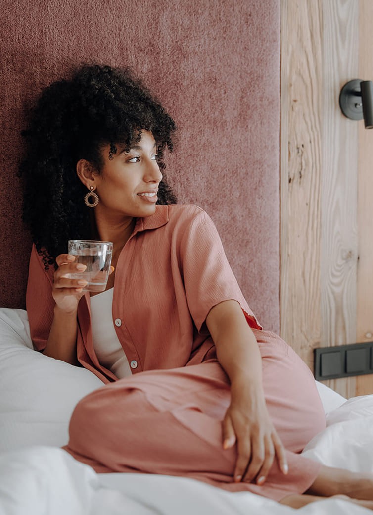 Frau in rosa Outfit sitzt entspannt im Bett und h&auml;lt ein Glas Wasser