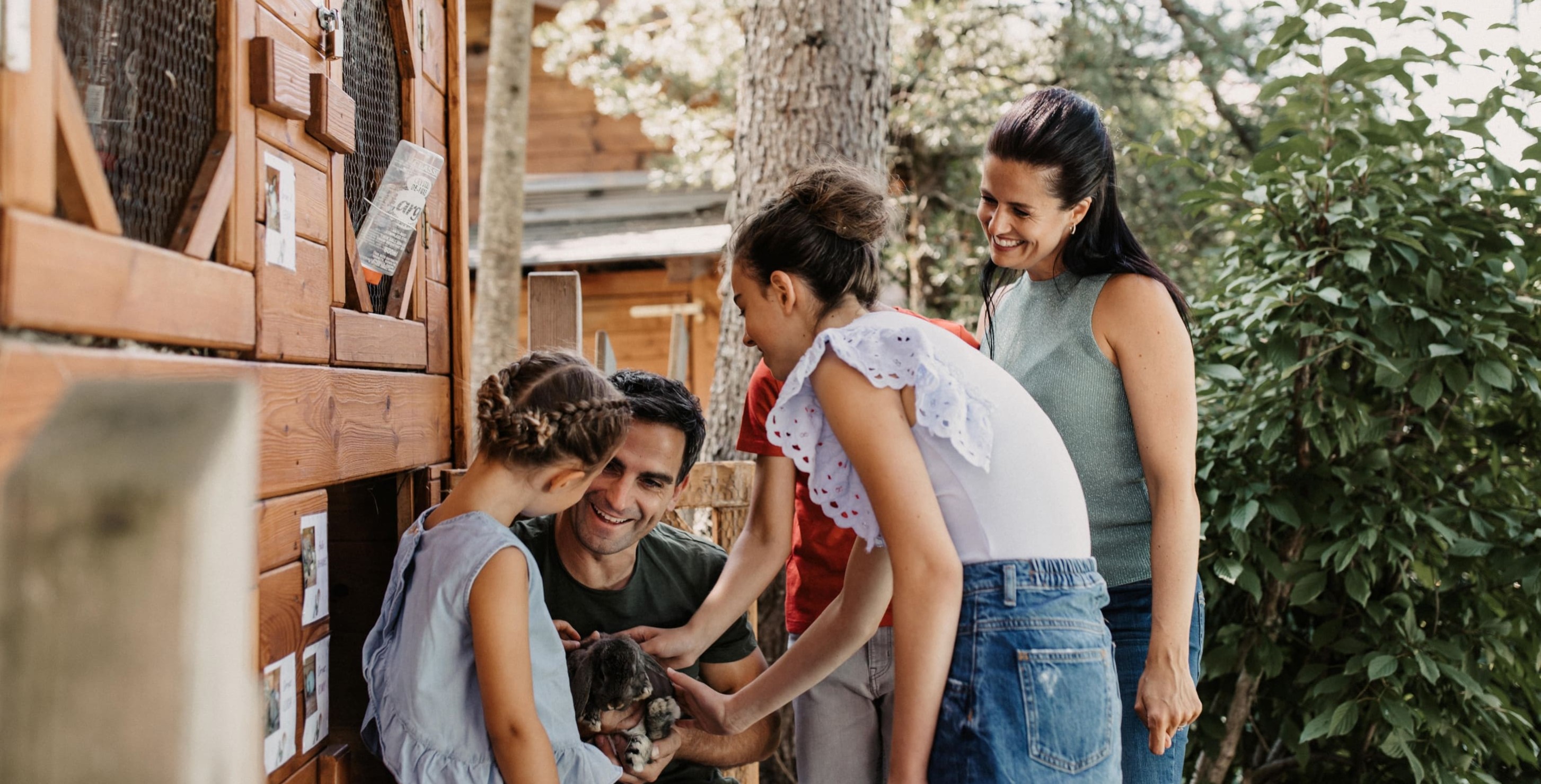 Familie streichelt Kaninchen vor einem Holzgehege im Grünen