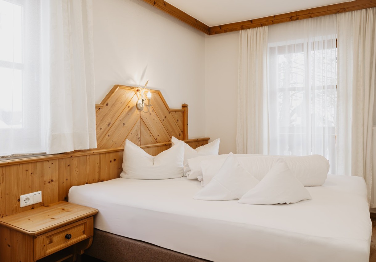 Helles Schlafzimmer mit Holzdekor und einem großen Bett vor zwei Fenstern mit weißen Vorhängen