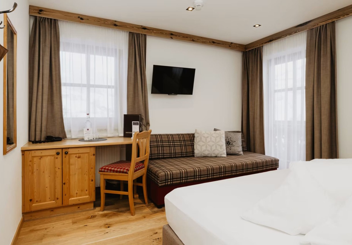 Helles Zimmer mit Holzmöbeln, Bett, Schreibtisch und Sofa vor großen Fenstern